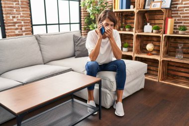 Young beautiful hispanic woman talking on smartphone with worried expression at home