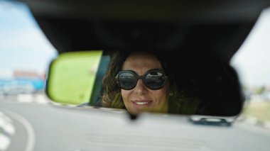 Orta yaşlı Latin bir kadın araba kullanıyor. Dikiz aynasından yola bakıyor.