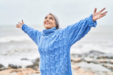 Orta yaşlı, kır saçlı, gülümseyen, kendine güvenen, deniz kenarında kolları açık duran bir kadın.