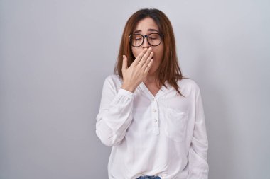 Esmer kadın izole edilmiş beyaz arka planda dikiliyor. Elle ağzını kapatmaktan sıkılmış. Huzursuz ve uykulu.. 