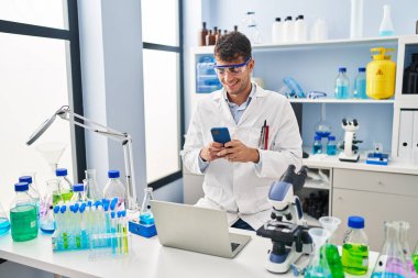 Young hispanic man scientist smiling confident using smartphone at laboratory