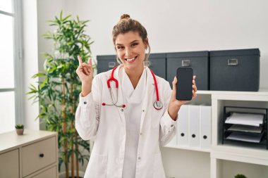Doktor üniforması giyen genç bir kadın elinde akıllı telefon, gülümsüyor. 