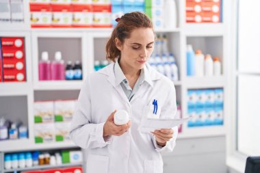 Young woman pharmacist holding pills bottle reading prescription at pharmacy
