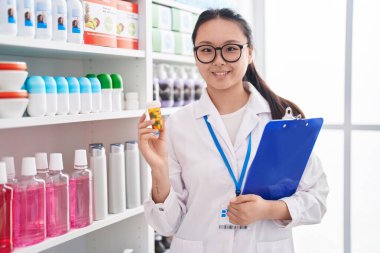 Genç Çinli kadın eczacı elinde haplarla eczanede doküman okuyor.