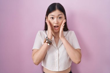 Chinese young woman standing over pink background afraid and shocked, surprise and amazed expression with hands on face 