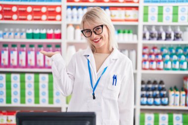 Young caucasian woman working at pharmacy drugstore smiling cheerful presenting and pointing with palm of hand looking at the camera. 