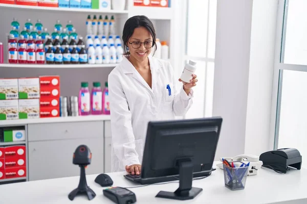 stock image Young hispanic woman pharmacist holding pills bottle using computer at pharmacy