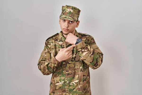 Young arab man wearing camouflage army uniform in hurry pointing to watch time, impatience, looking at the camera with relaxed expression