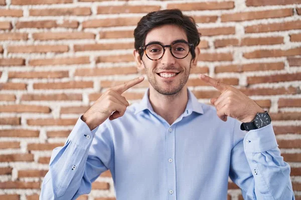 若いヒスパニック系の男がレンガの壁の背景に立って陽気に示す笑顔と指の歯と口で指しています 歯の健康の概念 — ストック写真