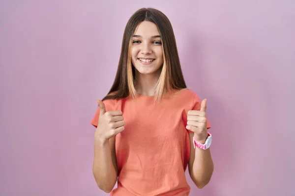 少女站在粉色背景的成功标志旁边 用手做着积极的手势 竖起大拇指 笑容满面 快乐无比 快乐的表达和胜利的姿态 — 图库照片