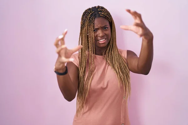 African american woman with braided hair standing over pink background shouting frustrated with rage, hands trying to strangle, yelling mad