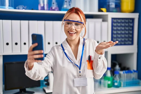 科学者研究室で働く若い白人女性が笑顔で達成を祝うスマートフォンでビデオ通話をし 手を挙げて勝者の表情 — ストック写真
