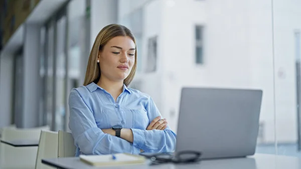 Young blonde woman business worker looking laptop with arms crossed gesture at office