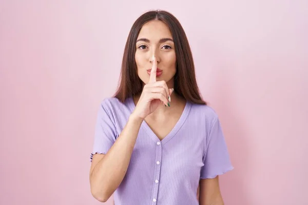 ピンクの背景に長髪の若いヒスパニック系の女性が唇に指で静かにするように求めて立っている 沈黙と秘密の概念 — ストック写真