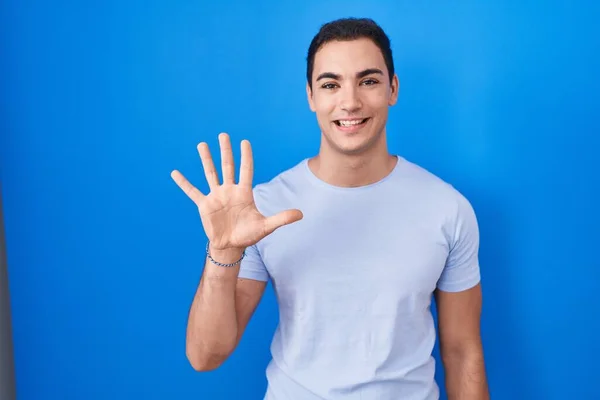 年轻的他的惊慌失措的男人站在蓝色的背景上 带着自信和快乐的微笑 用五号手指指指着 — 图库照片