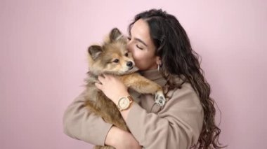 Köpeğiyle gülümseyen genç İspanyol kadın pembe arka planda duruyor.