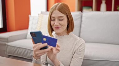 Akıllı telefon ve kredi kartıyla alışveriş yapan genç sarışın kadın evde yerde oturuyor.
