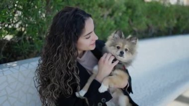 Köpeğiyle gülümseyen genç İspanyol kadın parkta bankta oturuyor.