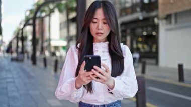 Young chinese woman using smartphone with serious expression at street