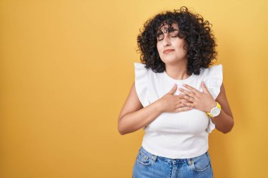 Orta Doğulu genç bir kadın sarı arka planda duruyor elleri göğsünde, gözleri kapalı ve yüzünde minnet dolu bir jest ile gülümsüyor. sağlık kavramı. 