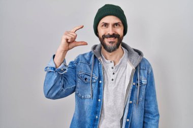 Yün şapkalı, gülümseyen, kendine güvenen, el hareketi yapan parmakları küçük gösteren ve kameraya bakan İspanyol dövmeli genç adam. ölçüm kavramı. 