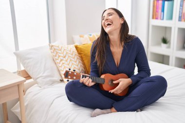 Young beautiful hispanic woman playing ukulele sitting on bed at bedroom