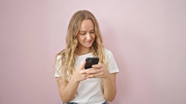 Akıllı telefon kullanan genç sarışın kadın pembe arka planda gülümsüyor.