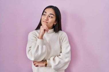 Pembe arka planda duran genç Güney Asyalı kadın, elleri çenesinde soru sorarken, düşünceli bir ifade takınıyor. Düşünceli bir yüzle gülümsüyor. şüphe kavramı. 