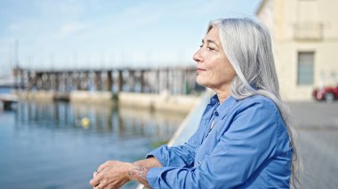 Orta yaşlı, kır saçlı bir kadın deniz kenarında ciddi bir ifadeyle yan tarafa bakıyor.