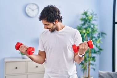 Young hispanic man smiling confident using dumbbells training at home