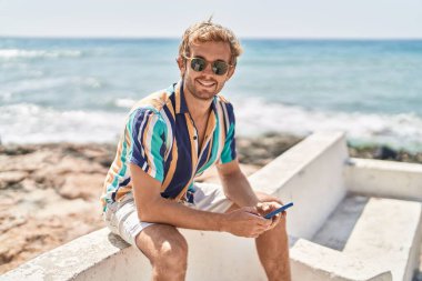 Young man tourist using smartphone sitting on bench at seaside