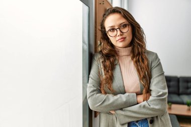 Genç, güzel, İspanyol bir iş kadını. Kollarını kavuşturup ofiste duruyor.