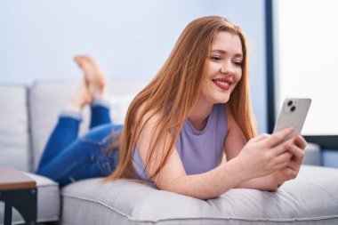 Young redhead woman using smartphone lying on sofa at home