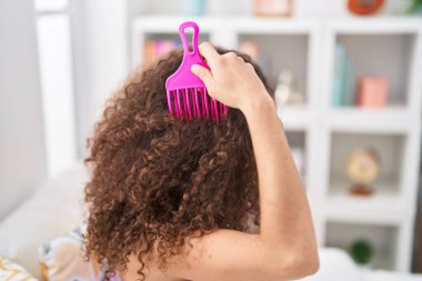 Young beautiful hispanic woman combing hair sitting on bed at bedroom