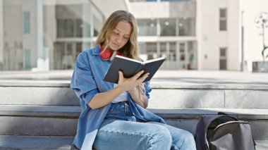 Üniversitede merdivenlerde oturmuş kitap okuyan genç sarışın kadın.