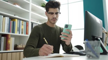 Young hispanic man student using smartphone writing on notebook at library university