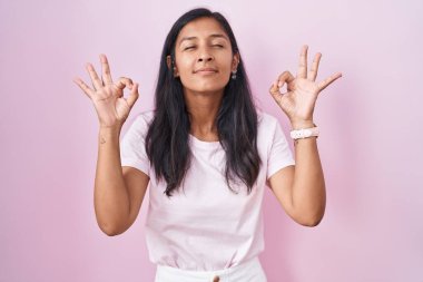 Young hispanic woman standing over pink background relaxed and smiling with eyes closed doing meditation gesture with fingers. yoga concept. 