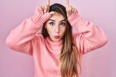 Young hispanic woman standing over pink background doing funny gesture with finger over head as bull horns 