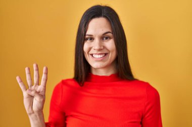 Young hispanic woman standing over yellow background showing and pointing up with fingers number four while smiling confident and happy. 