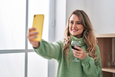Genç bir kadın akıllı telefonuyla selfie çekiyor. Evde şarap içiyor.
