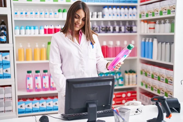 Young Beautiful Hispanic Woman Pharmacist Holding Shampoo Bottle Using Computer — Stok fotoğraf