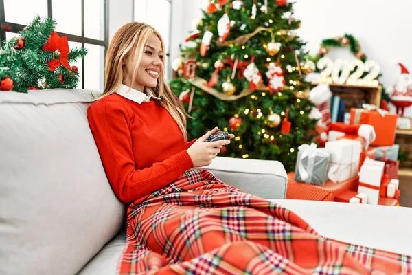 年轻的高加索女人在家里坐在圣诞树旁的沙发上玩电子游戏 — 图库照片