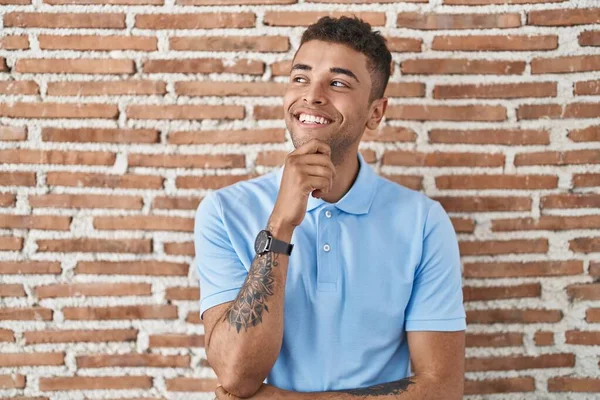集中的な表現について考える顎の手でレンガの壁の上に立っているブラジルの若い男 笑顔で思いやりのある顔 疑わしい概念 — ストック写真