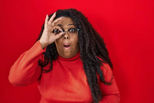 stock image Plus size hispanic woman standing over red background doing ok gesture shocked with surprised face, eye looking through fingers. unbelieving expression. 