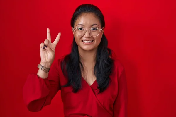 亚洲的年轻女子站在红色的背景上 微笑着 自信而快乐 同时又用第2号手指指指着 — 图库照片