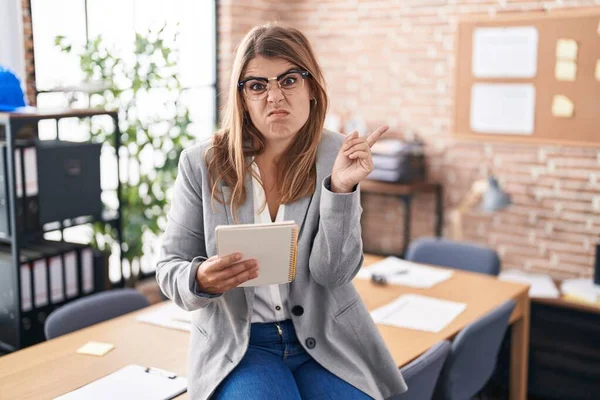 Young hispanic woman working at the office wearing glasses pointing aside worried and nervous with forefinger, concerned and surprised expression