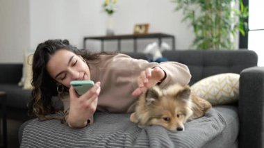Köpekli genç İspanyol kadın evdeki kanepede uzanmış akıllı telefonuyla sesli mesaj yolluyor.