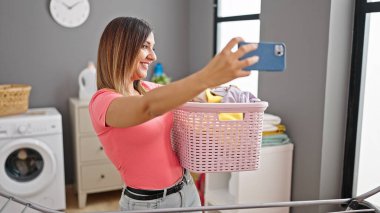 Orta Doğulu bir kadın çamaşır odasında selfie çekerken elinde sepetle gülümsüyor.