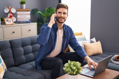 Young caucasian man talking on smartphone using laptop at home