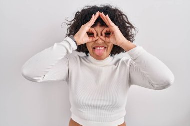 Hispanic woman with curly hair standing over isolated background doing ok gesture like binoculars sticking tongue out, eyes looking through fingers. crazy expression. 